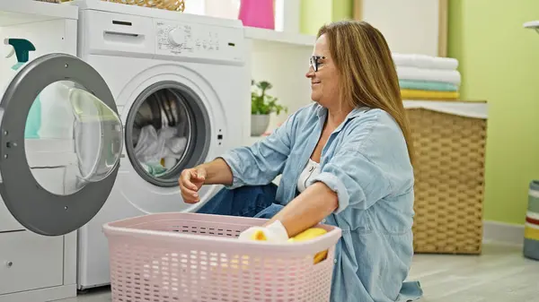 Middle age hispanic woman washing clothes sitting on floor at laundry room