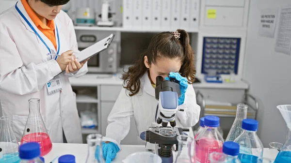 Two serious science girls, partners working together under microscope, diligently taking notes in lab room indoors. professional experiment in medical analysis and research.