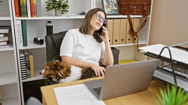 A professional young woman talks on the phone while petting her dog at her organized office space.