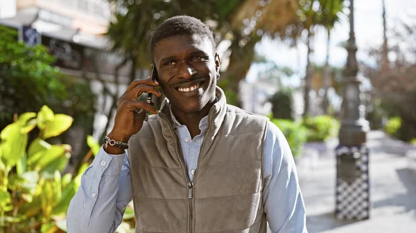 Smiling african man making a phone call outdoors in a sunny city park.