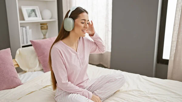 Smiling woman in pajamas enjoying music with headphones while sitting on a bed in a cozy bedroom