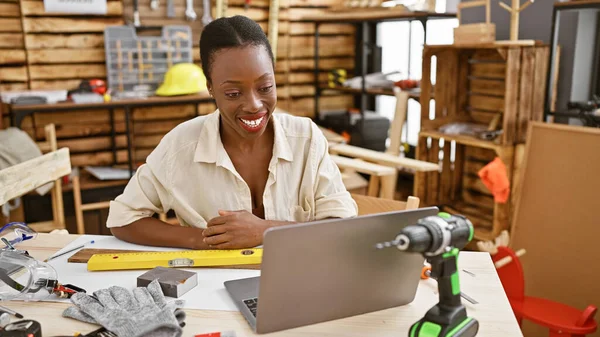 Beautiful african american carpenter woman smiling as she works on her woodworking business with laptop at carpentry workshop