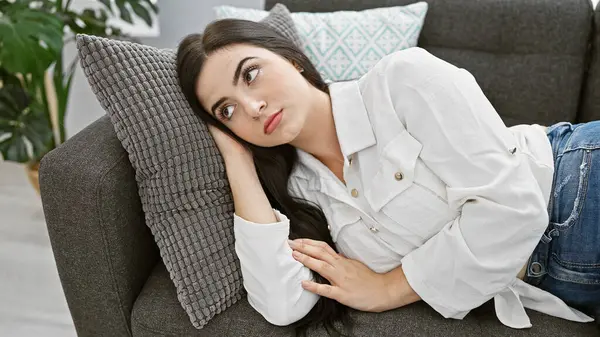 Düşünceli, genç İspanyol bir kadın, evin içinde bir kanepede uzanıyor, rahat bir evde güzellik ve zarafet yansıtıyor..