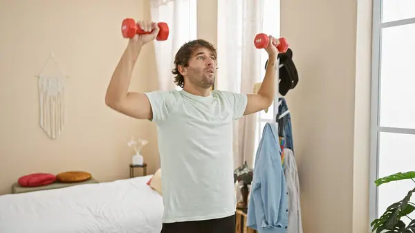 A young man lifts weights in a well-lit bedroom, showcasing fitness and a casual home workout environment.