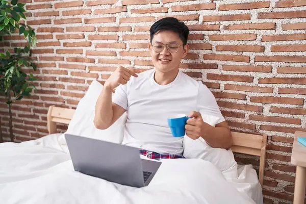 Young asian man drinking coffee sitting on the bed pointing finger to one self smiling happy and proud