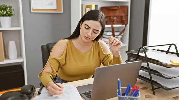 stock image A focused hispanic woman takes notes in an office setting, highlighting business, workplace, and professional themes.