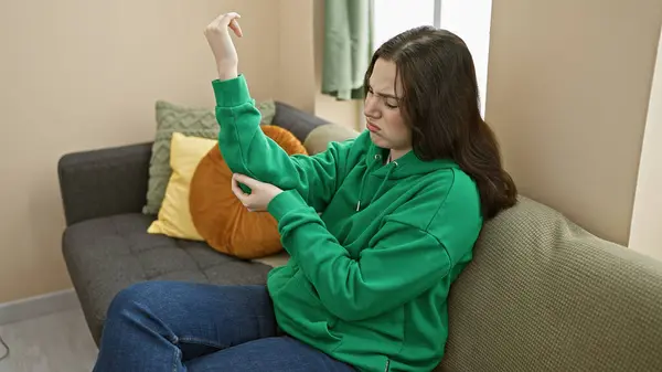 一位穿着绿色帽衫的年轻女子坐在灰色的沙发上 坐在室内检查她的肘部 看上去很担心 — 图库照片