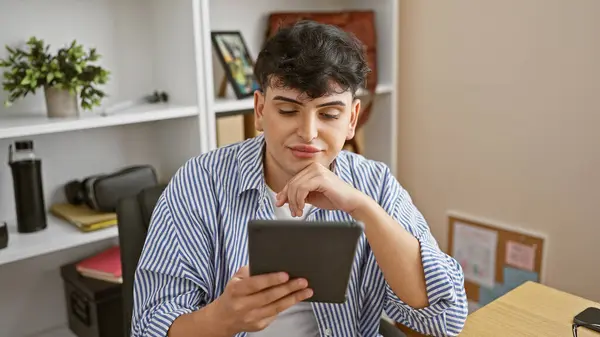 A young man dressed in a striped shirt sits in an office, intently reading something on his tablet.
