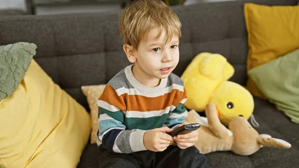 金发蹒跚学步的男孩坐在室内沙发上 在舒适的客厅里玩遥控器和毛绒玩具 — 图库照片