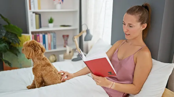 Young caucasian woman with dog reading book sitting on bed at bedroom
