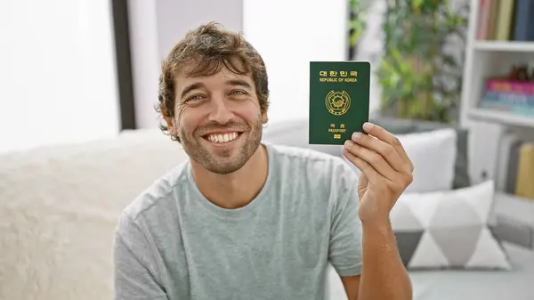 Young man sitting on sofa holding passport of korea smiling at home