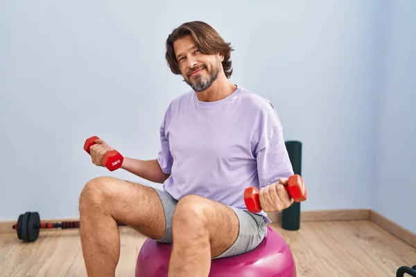 Middle age man training with dumbbells sitting on fit ball at sport center