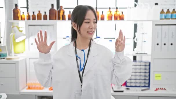 幸せな若い中国人女性科学者は 研究室で白い実験室のコートを着て 明るく微笑んで指で8番を示しています — ストック動画