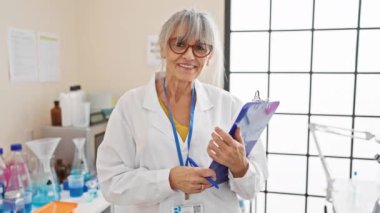 Gri saçlı, gözlüklü ve laboratuvar ortamında elinde bir pano tutan gülümseyen olgun kadın bilim adamı..