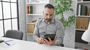 Gri sakallı, olgun iş adamı modern ofiste tablet kullanıyor.