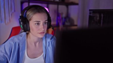 Genç, çekici, kulaklık takan sarışın bir kadın geceleri renkli, karanlık bir oyun odasında perdeye odaklanıyor..