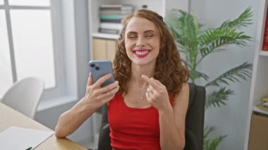 Kendine güvenen ve ışıl ışıl genç kadın gülümseyen yüzüyle ofisin içinde akıllı telefonunu kullanıyor. Neşeli ifade heyecanlı bir şey yakaladığını gösteriyor.! 