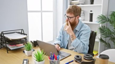 Kendine güvenen genç kızıl saçlı adam ofisteki laptopta neşeli bir şekilde çalışıyor, iş adamı olarak kendine güveniyor.