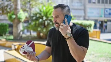 Kızgın, olgun, sakallı, sakallı bir adam, elinde hediye tutarken telefonla konuşuyor.