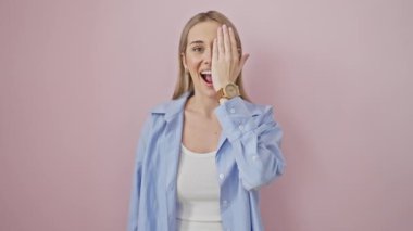 Neşeli, genç, sarışın bir kadın. Bir gözünü eliyle kapatıyor. Pembe izole edilmiş arka planda kendine güvenen bir gülümseme sergiliyor.