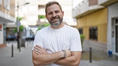 Gri saçlı ve sakallı olgun bir adam şehir ortamında güvenle gülümsüyor..