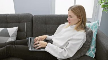 Stresli genç sarışın bir kadın, rahat bir odadaki kanepede dizüstü bilgisayar kullanıyor ve yüzünü yastıkla örtüyor..