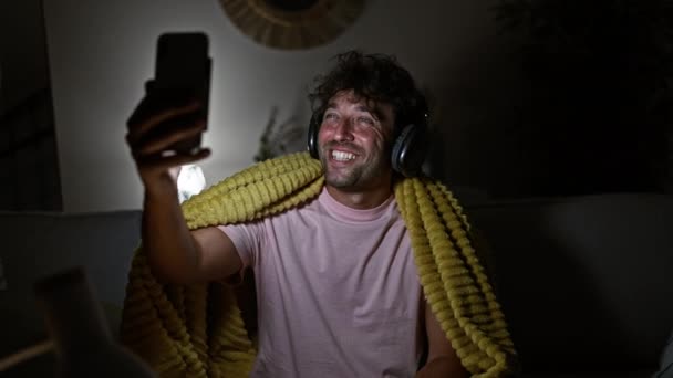 Glimlachende Man Met Koptelefoon Neemt Selfie Thuis Tijdens Avond — Stockvideo