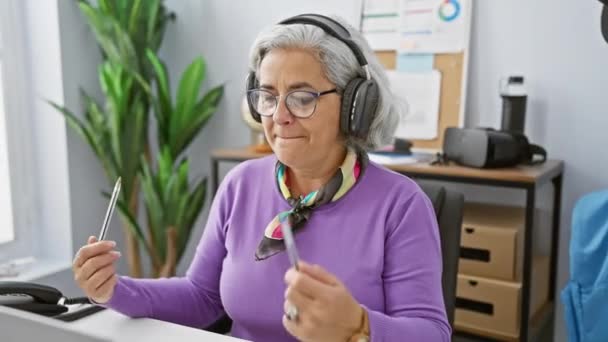 オフィスで働く灰色の髪を持つ成熟した女性 ペンを保持し ラップトップの画面を見ながらヘッドフォンや笑顔を身に着けています — ストック動画
