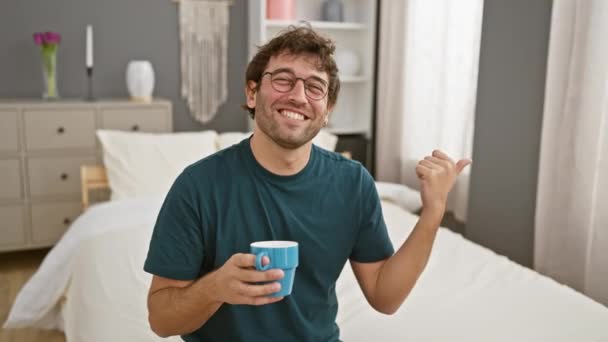陽気な若いヒスパニックの男性は 自信を持ってベッドにコーヒーを飲んで 親指のジェスチャーを指差し 家のインテリアの中で居心地の良い部屋で彼の友好的で楽しい笑顔を明らかに彼の口 — ストック動画