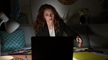 Genç bir kadın, dokümanlar ve dizüstü bilgisayarla çevrili bir şekilde gece geç saatlere kadar çalışırken ceketini çıkarıyor..