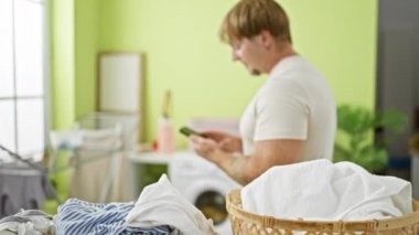 Açık yeşil bir odada çamaşır yıkarken akıllı telefon kullanan sakallı genç bir adam..