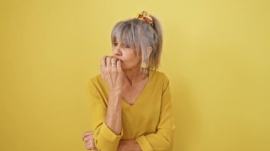 Gergin, orta yaşlı, gri saçlı, tırnak yiyen bir kadın. At kuyruğu takan olgun bir kadın sarı arka planda izole edilmiş stres ifadesi gösteriyor.