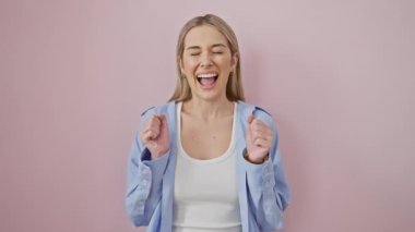 Coşkulu kazanan! Gömlekli genç sarışın kadın, kollarını muzaffer bir eğlence için kaldırdı! Gözler kapalı, pembe arka planda soyutlanmış, başarıyı neşeli bir gülümsemeyle kutluyor..