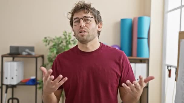 Ung Hispanisk Mann Med Briller Skjeggbevegelser Mens Han Snakker Innendørs – stockvideo