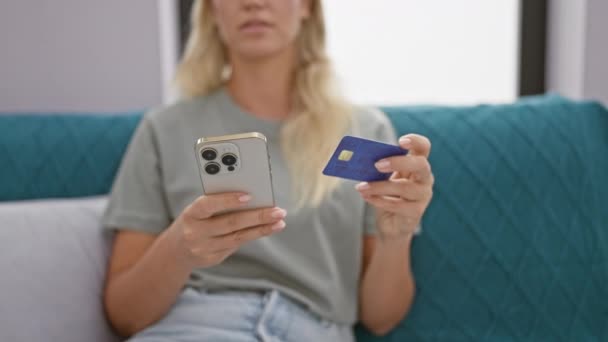 ペンシルベニアの若い女性は 現代のリビングルームでスマートフォンを保持しながらクレジットカードを調べます — ストック動画