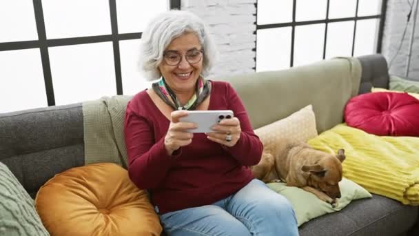 居心地の良いリビングルームでペット犬とソファーでスマートフォンを使用して幸せな高齢女性 — ストック動画