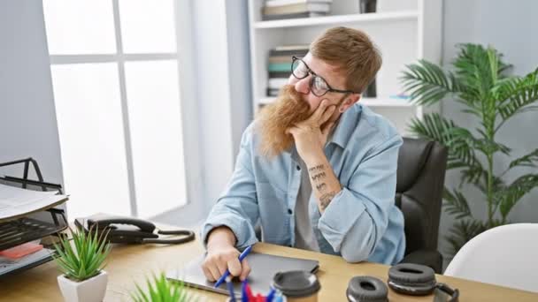 一个严肃的红头发年轻人在怀疑一个想法 一个优雅的生意人正在工作场所的室内打坐 办公室里拿着笔记本电脑坐在桌旁 — 图库视频影像