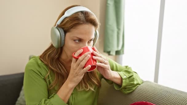 身穿绿色衬衫的西班牙裔女性在家里一边欣赏着咖啡 一边用白色耳机听音乐 — 图库视频影像