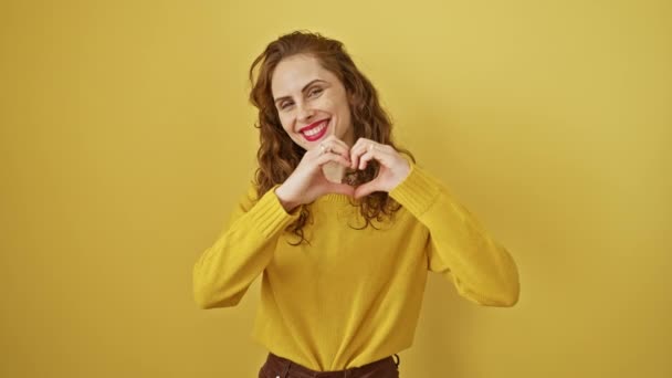 黄色い背景に隔離された放射性の若い女性は 手で愛の心を形作ります ロマンチックな幸せの美しいシンボル 笑顔のジェスチャーで描かれた陽気なバレンタインのコンセプト — ストック動画