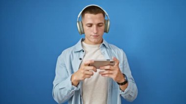 Kulaklıklı genç bir adam mavi duvara karşı akıllı telefon kullanıyor. Bağlılık ve konsantrasyonu ifade ediyor..