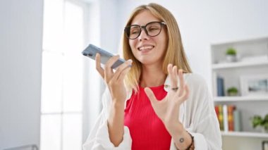 Ofiste genç bir kadın, gözlük, beyaz ceket ve kırmızı bluz giyerken işaret eden bir akıllı telefona konuşur..