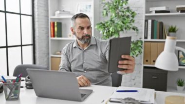 Olgun sakallı bir adam modern ofis ortamında özçekim yapıyor. Tabletini kullanırken hayal kırıklığı ve odaklanma gösteriyor..