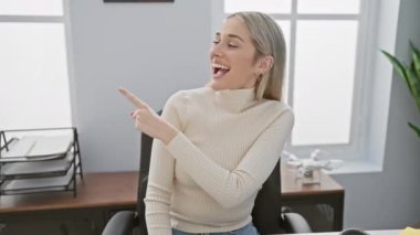 Ofis masasında oturan neşeli, genç sarışın kadın, mutlu bir şekilde kendinden emin bir ifadeyle yüzünü yana çeviriyor ve gülümsüyor.