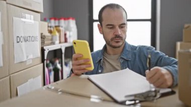 Odaklanmış, kel, sakallı, akıllı telefon kullanırken bir odada bağış organize eden bir İspanyol..