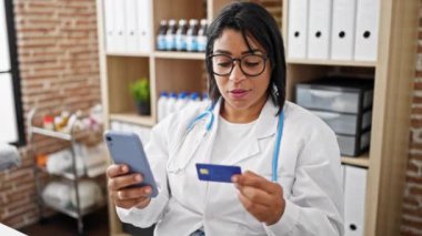 Beyaz laboratuvar önlüklü, gülümseyen bir İspanyol kadın. Kliniğin iç mekânında akıllı telefon ve kredi kartı kullanıyor.