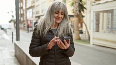 Şehir caddesinde akıllı telefon kullanan gri saçlı olgun bir kadın, günlük yaşamında teknolojiyi somutlaştırıyor..