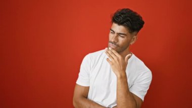 Sakallı yakışıklı, genç, İspanyol bir adam izole edilmiş bir kırmızı duvara karşı düşünceli bir şekilde poz veriyor. Bir anlık tefekkür ya da fikir kuşağını somutlaştırıyor..