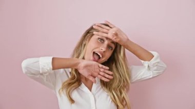 Neşeli genç sarışın kadın dikizleme oyunu oynuyor! Pembe, soyutlanmış bir arka planda, tişört giyerken sevinçle şaşırmış yüzünü gösteriyor..