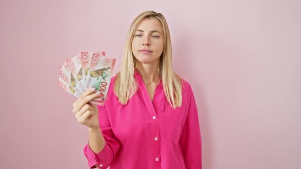 陽気な若いブロンドの女性は誇らしげに新しいゼーランドドルを表示し 楽観的な笑顔を指し 孤立したピンクの背景の上に — ストック動画