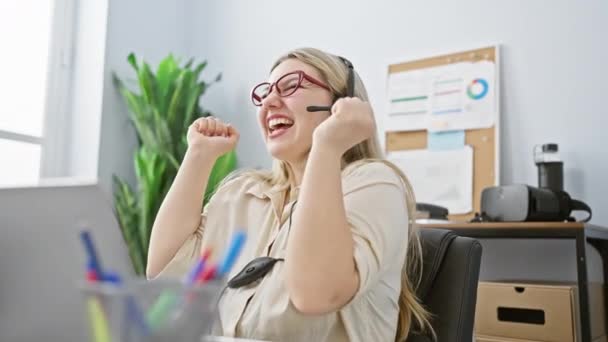 メガネとヘッドセットを持つ陽気な若い女性は 仕事で喜びの瞬間を経験し オフィスデスクで成功を祝います — ストック動画